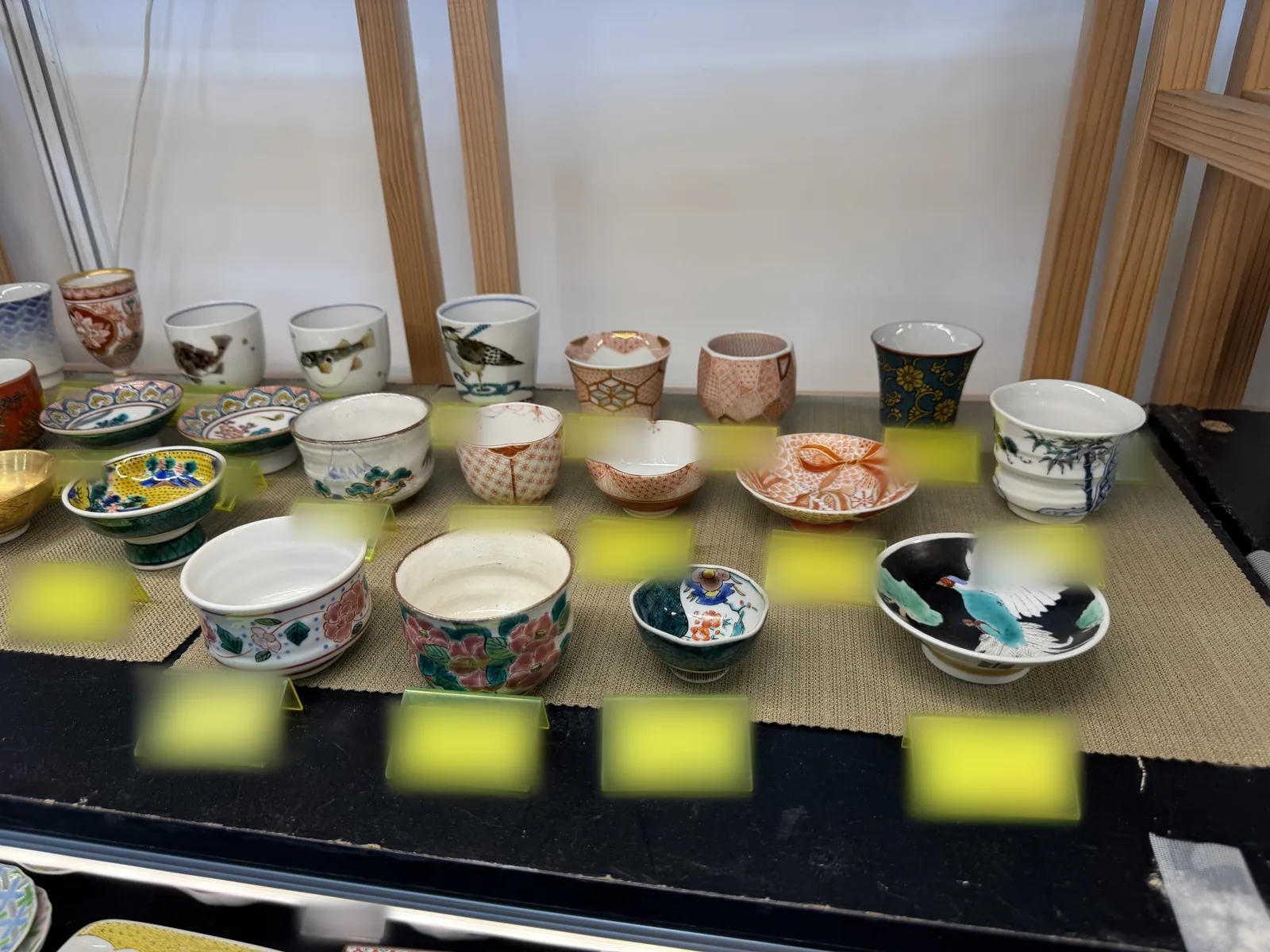 A shelf of Kutani ware cups, bowls, and sake cups on a woven mat at the booth, with the price tags blurred