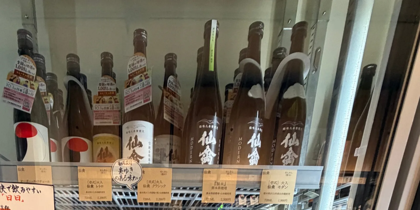 A shelf of Senkin bottles with handwritten tags in the refrigerated case at Inomoto Saketen in Kumamoto